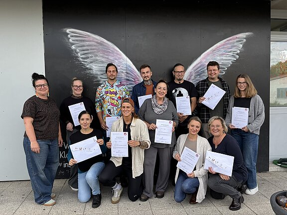 Eine Gruppe von Menschen posiert vor einer Wand mit gemalten Engelsflügeln und hält Zertifikate in den Händen.